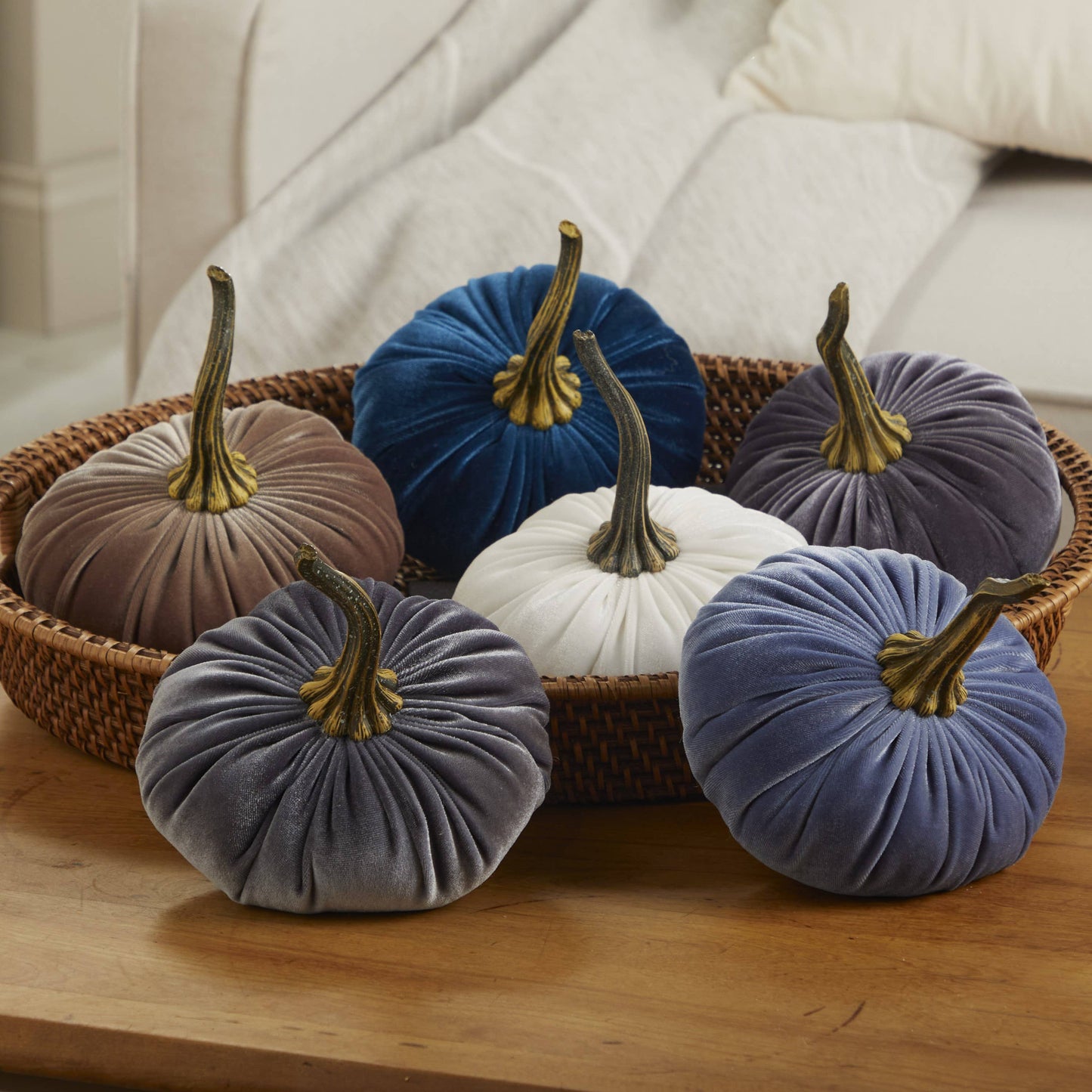 A wooden table displays a wicker tray holding six “Your Heart’s Content - Pumpkin Large Velvet” decorations by Your Heart’s Content in blue, gray, white, brown, and purple, each featuring a gold stem—ideal as handcrafted fall mantel decor or a unique Thanksgiving centerpiece. A cream sofa is visible in the background.