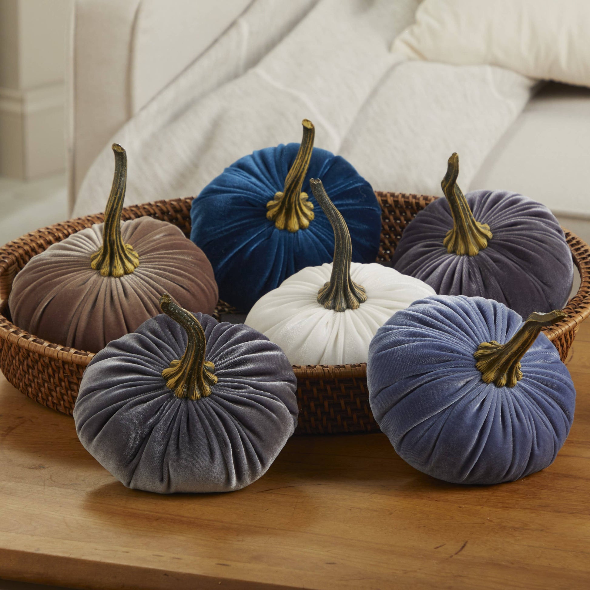 A wooden table displays a wicker tray holding six “Your Heart’s Content - Pumpkin Large Velvet” decorations by Your Heart’s Content in blue, gray, white, brown, and purple, each featuring a gold stem—ideal as handcrafted fall mantel decor or a unique Thanksgiving centerpiece. A cream sofa is visible in the background.