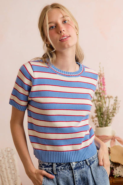 Wearing the So Me - STRIPE KNIT CREW NECK SHORT SLEEVES SWEATER TOP, featuring blue and white stripes with red accents, a person stands against a soft pink backdrop adorned with flowers and plants, capturing the effortless charm of So Me style.