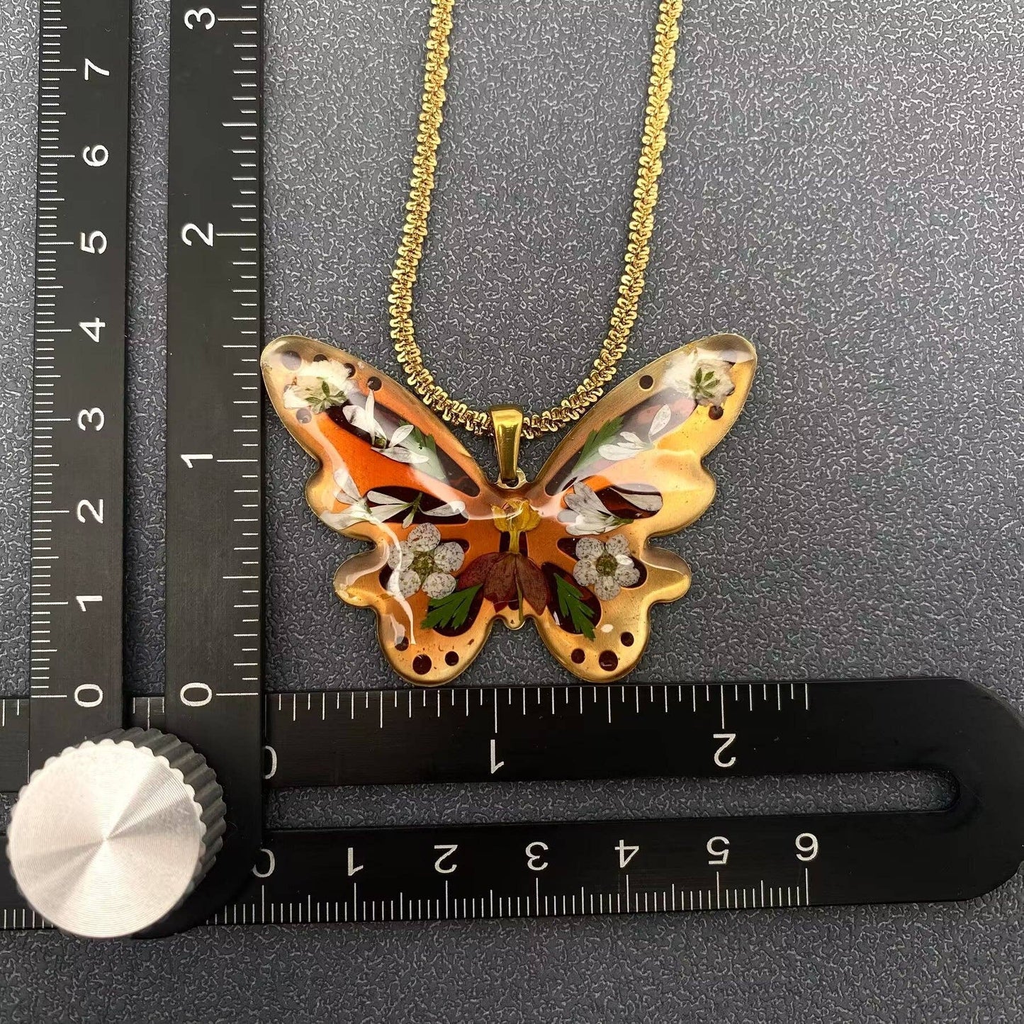 A "Mio Queena - Dried Flower Brass Butterfly 18K Golden SS Necklace - PDF" by Mio Queena, featuring a butterfly-shaped pendant with colorful dried flower designs, is suspended from a gold plated chain and placed at the intersection of two black rulers displaying centimeter and inch measurements.