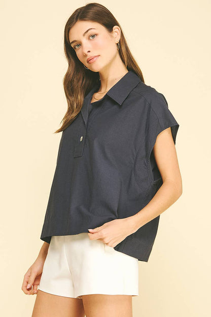 A woman with long brown hair wears the PINCH SOLID CAP SLEEVE BLOUSE TOP, a loose, classic-collared navy blouse by PINCH, paired with white shorts and standing against a beige background.