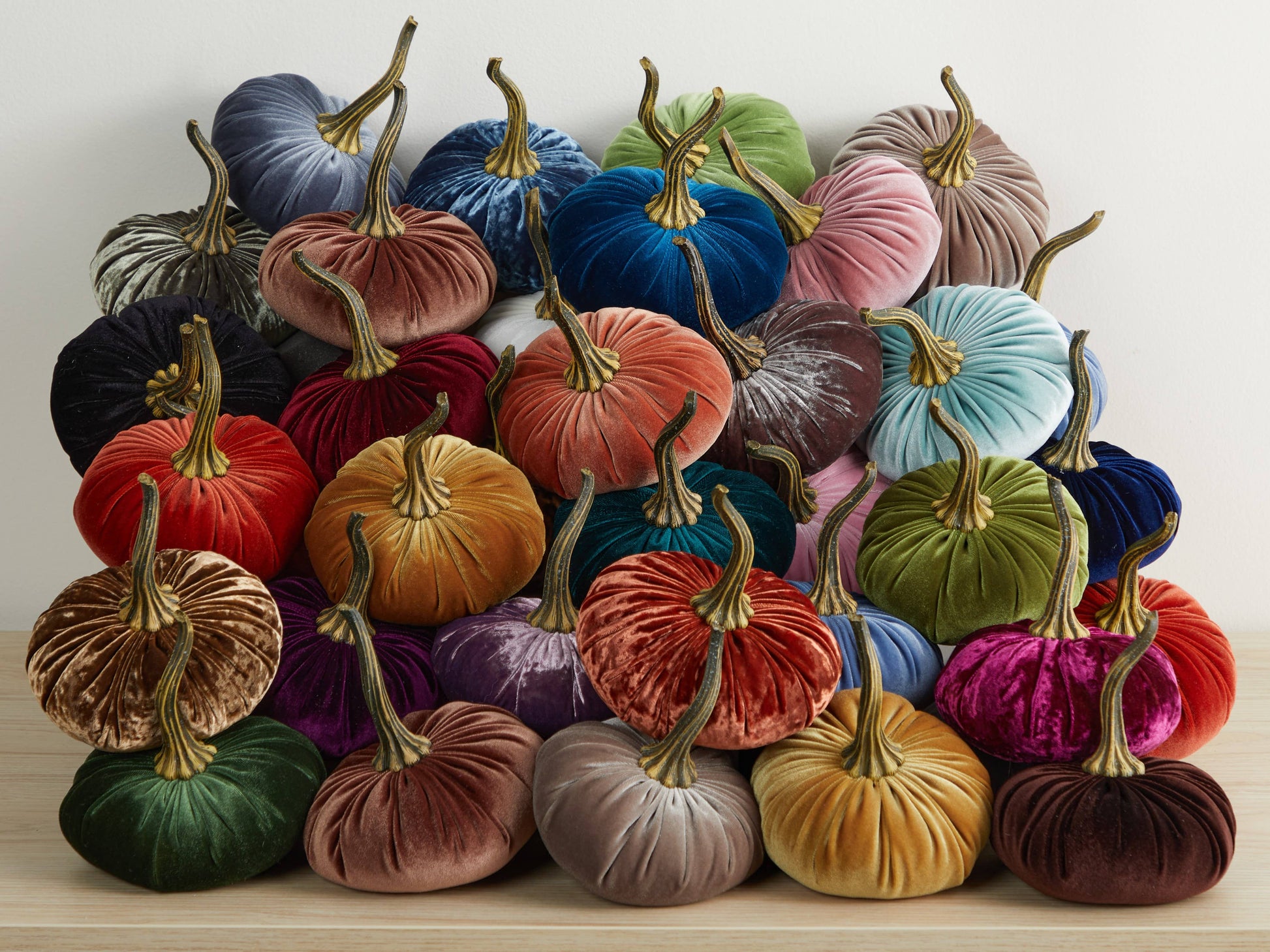 A stack of Your Heart’s Content - Pumpkin Large Velvet pumpkins in a range of vibrant colors rests on a wooden surface against a plain white wall, creating a striking handcrafted arrangement from Your Heart’s Content that’s perfect for Thanksgiving centerpieces, fall mantel decor, or Halloween tables.