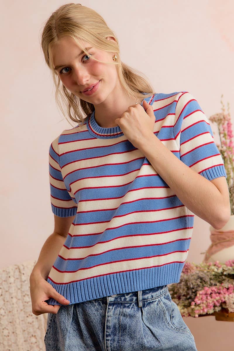 A woman is seen indoors, smiling against a light pink background with flowers behind her, wearing the So Me STRIPE KNIT CREW NECK SHORT SLEEVES SWEATER TOP in blue and red, styled with high-waisted jeans.