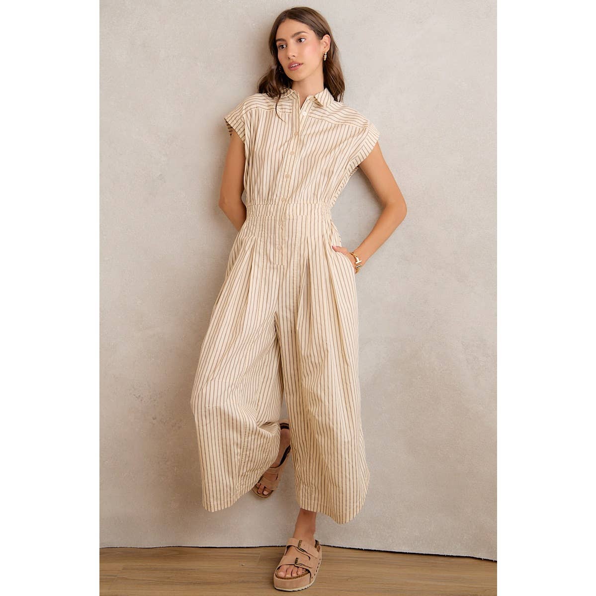 A woman stands against a light-colored wall with one leg slightly bent and hands in the pockets of the So Me SLEEVELESS WIDE LEG STRIPE JUMPSUIT, featuring beige and white stripes, styled with sandals.