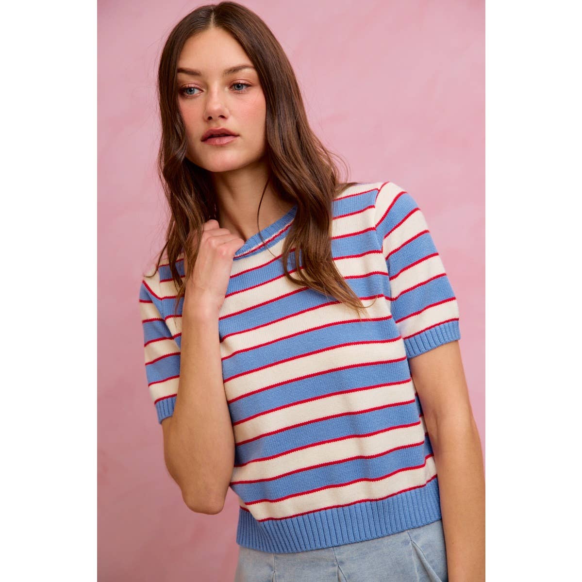 A young woman with long brown hair wears the So Me - STRIPE KNIT CREW NECK SHORT SLEEVES SWEATER TOP from So Me, featuring a stylish design with blue and white stripes accented by red lines. She stands against a pink background, gazing off to the side with a neutral expression.
