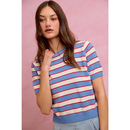 A young woman with long brown hair wears the So Me - STRIPE KNIT CREW NECK SHORT SLEEVES SWEATER TOP from So Me, featuring a stylish design with blue and white stripes accented by red lines. She stands against a pink background, gazing off to the side with a neutral expression.