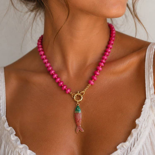 A woman wearing a white top and the vibrant JOSSLYN by wall to wall Stainless Steel Tarnish Free Fish Beaded Necklace gazes off to the side. The image highlights the necklace's lasting shine and beauty, drawing attention to its gold fish pendant and her neckline.