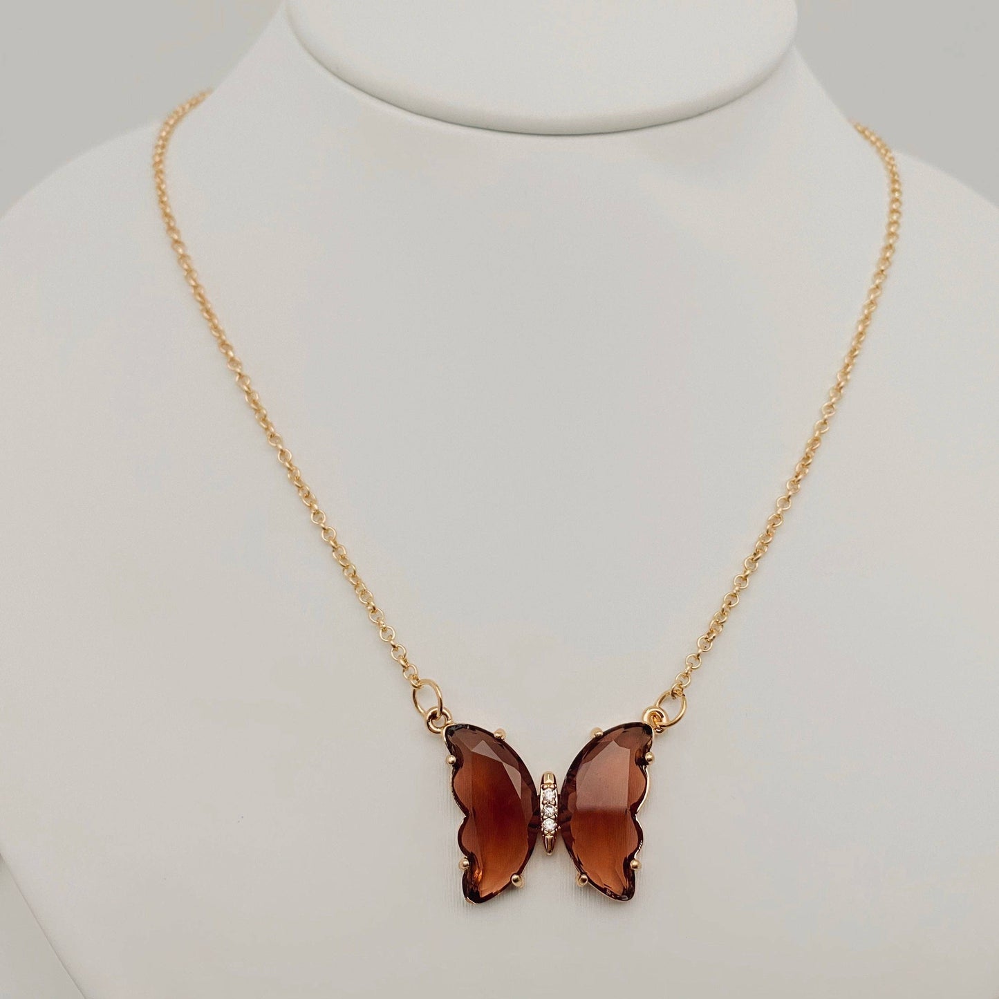 The Mio Queena - Multicolor Glass Butterfly Pendant Necklace by Mio Queena features a butterfly-shaped glass pendant in translucent brown, suspended from a gold chain and elegantly displayed on a white jewelry stand.