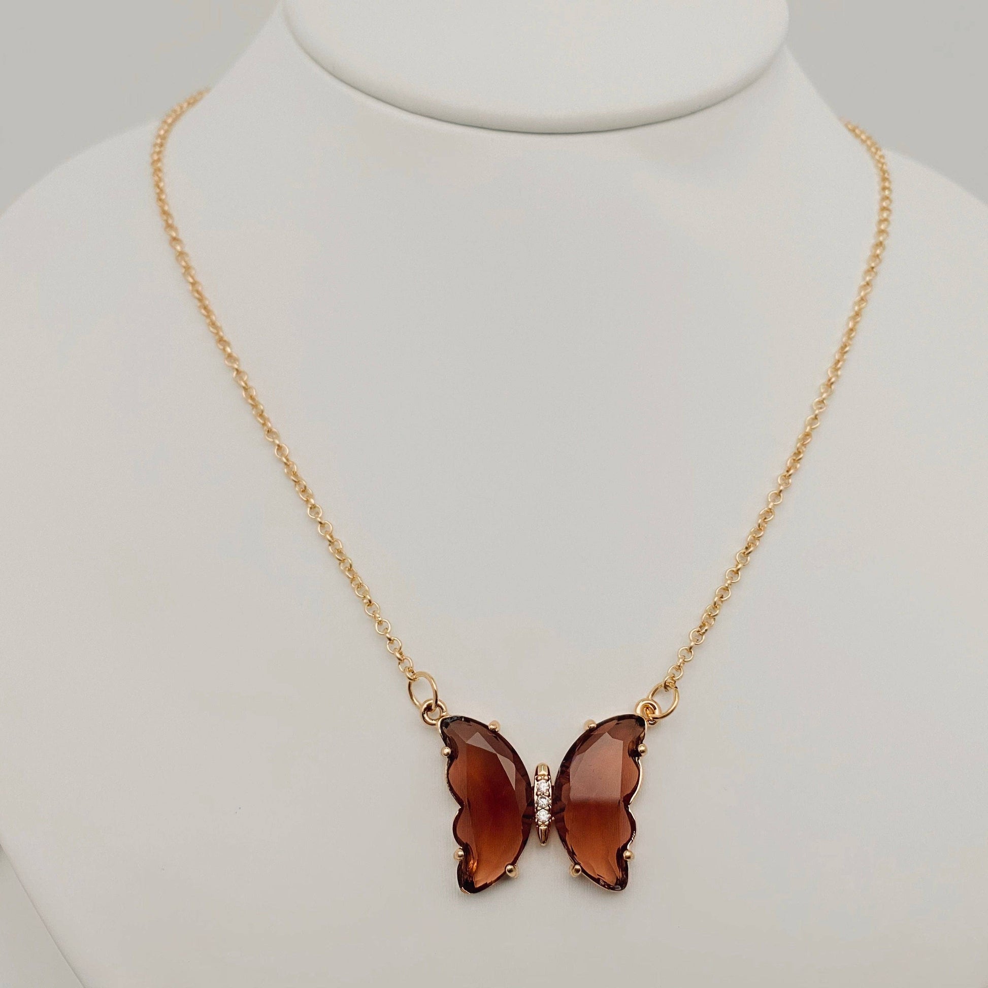 The Mio Queena - Multicolor Glass Butterfly Pendant Necklace by Mio Queena features a butterfly-shaped glass pendant in translucent brown, suspended from a gold chain and elegantly displayed on a white jewelry stand.