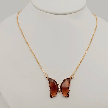 The Mio Queena - Multicolor Glass Butterfly Pendant Necklace by Mio Queena features a butterfly-shaped glass pendant in translucent brown, suspended from a gold chain and elegantly displayed on a white jewelry stand.