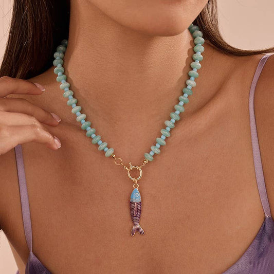A woman is shown from shoulders to chin wearing the JOSSLYN by wall to wall Stainless Steel Tarnish Free Fish Beaded Necklace—a light blue beaded necklace featuring a gold clasp and a colorful fish pendant. This stylish piece perfectly complements her lavender spaghetti strap top.