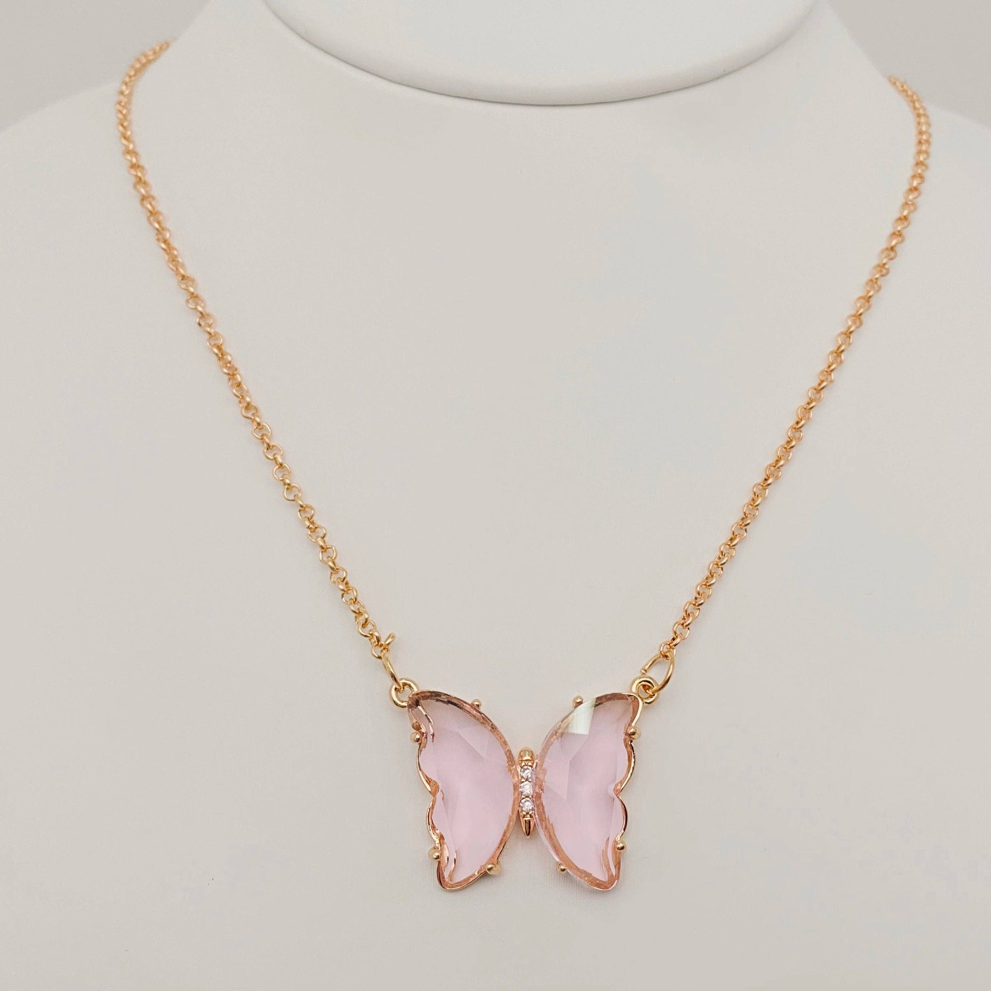 The Mio Queena - Multicolor Glass Butterfly Pendant Necklace by Mio Queena features a gold butterfly-shaped pendant with translucent pink glass wings and a delicate row of clear stones at the center, displayed on a white stand. This beautiful piece is also hypoallergenic, making it comfortable for everyday wear.
