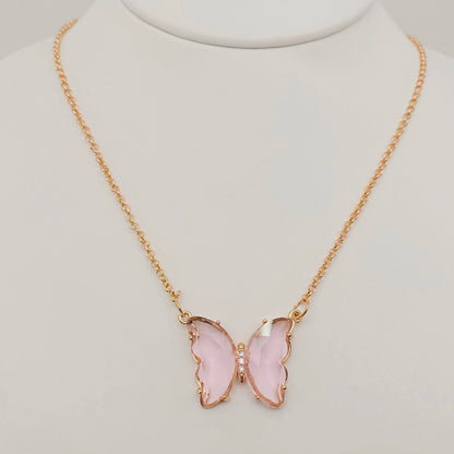 The Mio Queena - Multicolor Glass Butterfly Pendant Necklace by Mio Queena features a gold butterfly-shaped pendant with translucent pink glass wings and a delicate row of clear stones at the center, displayed on a white stand. This beautiful piece is also hypoallergenic, making it comfortable for everyday wear.