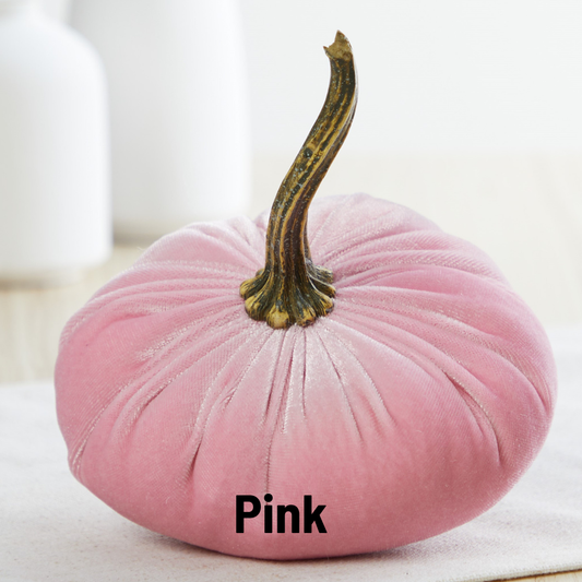 A small velvet pumpkin from Your Heart’s Content, in a soft pink hue with a brown stem, sits on a white surface—perfect for adding charm to your Valentine’s mantel or autumn table. The word “Pink” appears at the base of the pumpkin, while two blurred white objects are visible in the background.