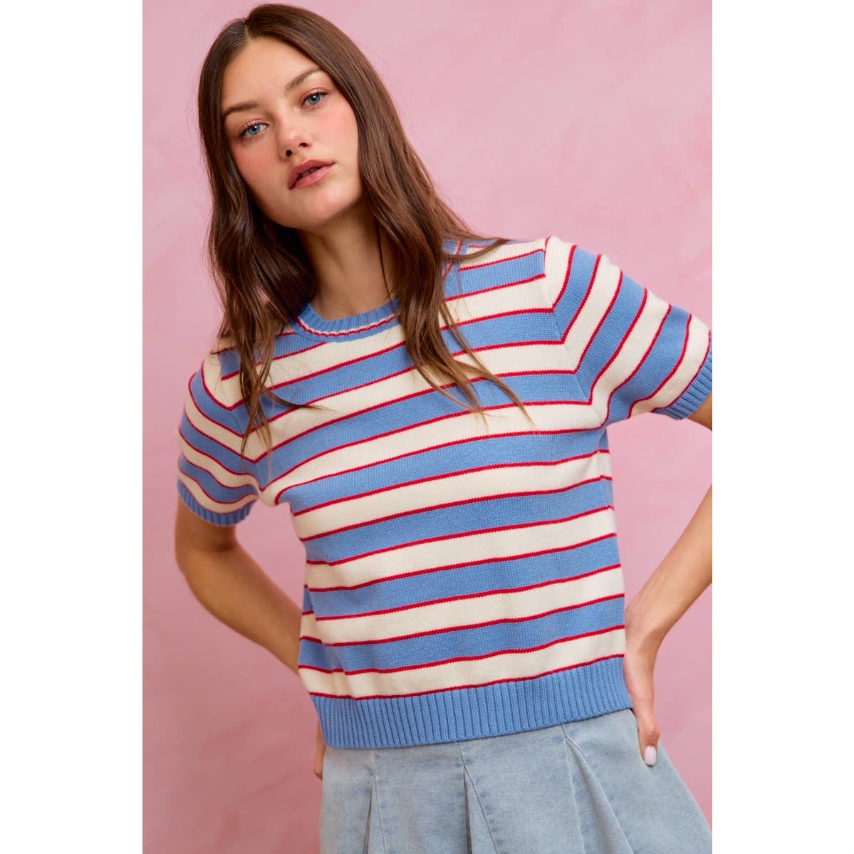 A woman with long brown hair is wearing the So Me - STRIPE KNIT CREW NECK SHORT SLEEVES SWEATER TOP by So Me, featuring white, blue, and red stripes, paired with a light blue skirt as she stands against a pink background.