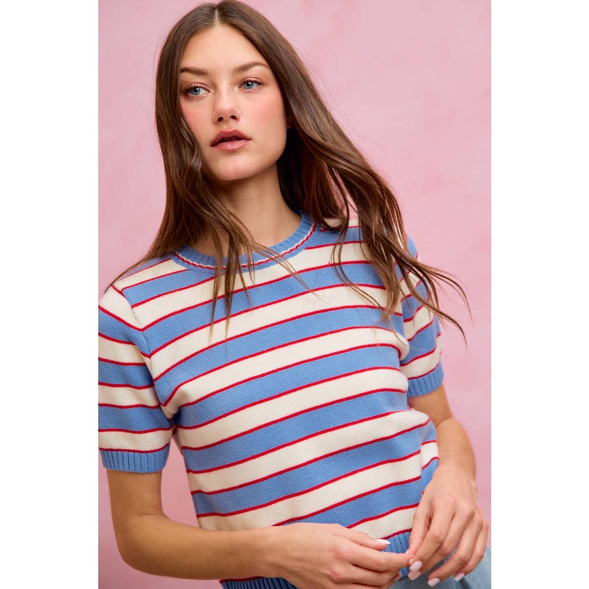 A woman with long brown hair stands in front of a pink background wearing the So Me STRIPE KNIT CREW NECK SHORT SLEEVES SWEATER TOP, which features blue and white stripes with red accents.