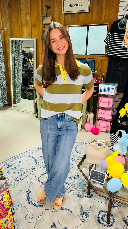 A young woman with long brown hair smiles as she wears the &MERCI Striped Sweater Knit Short Sleeve Polo and wide-leg jeans, standing on a patterned rug in a cozy, colorful boutique filled with boxes and decor items.