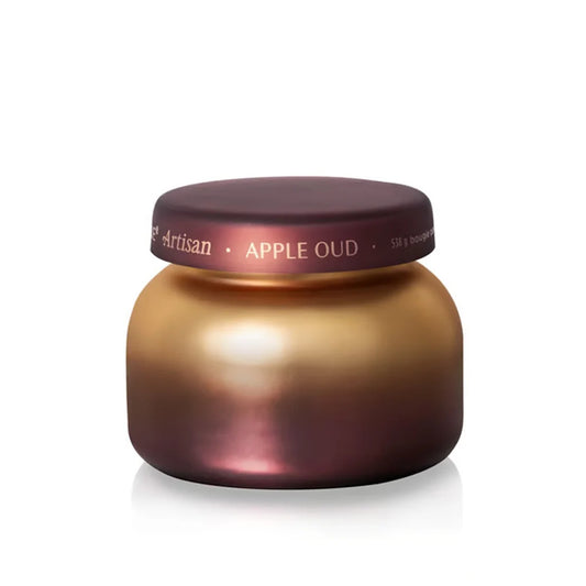 A small, round jar with a metallic gold and burgundy ombré gradient finish, labeled “Capri Blue Apple Oud Radiance Signature Candle” on the lid, displayed against a white background—this elegant candle by Capri Blue is the perfect luxury home fragrance accent and a stunning piece of ombré jar decor.