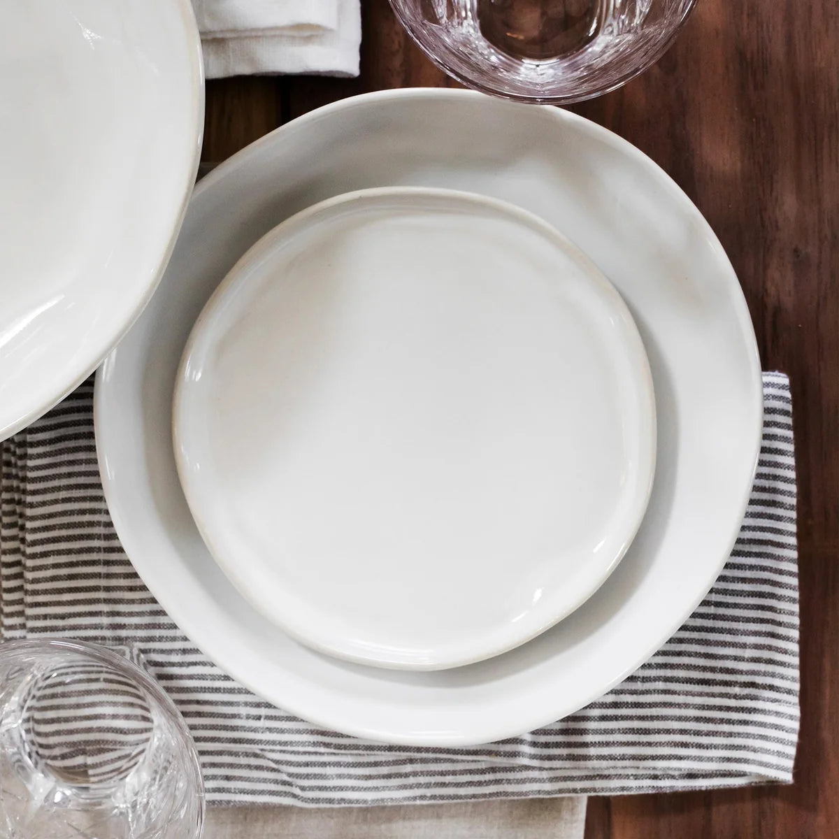 Two stacked Vietri “Forma Cloud” salad plates rest on a striped cloth napkin atop a wooden table, with clear glassware nearby.