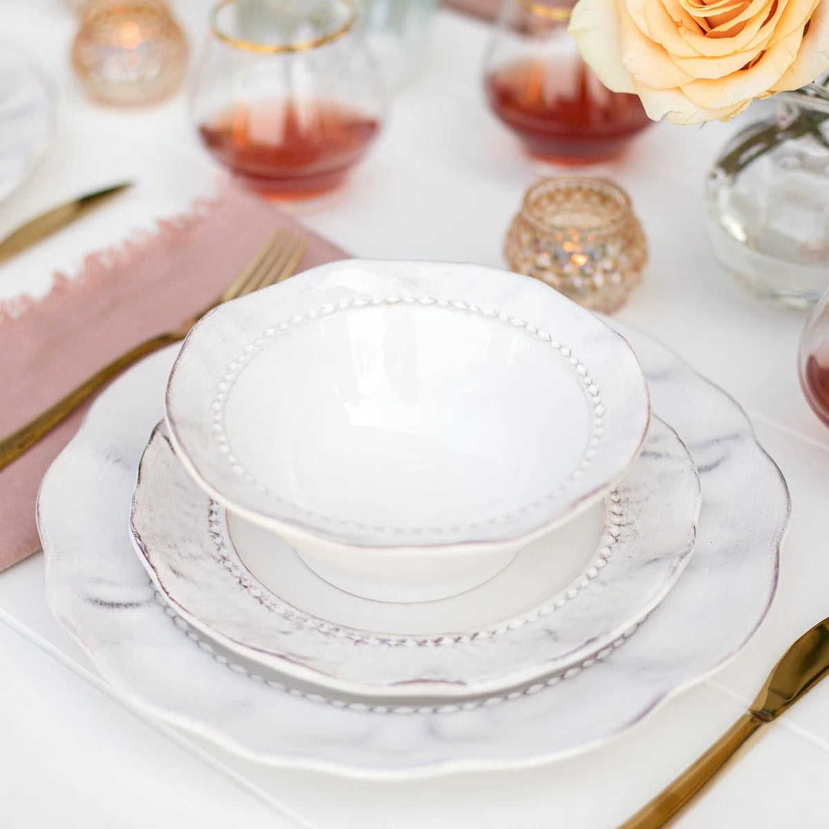 Elegant table setting featuring the Vietri “Giorno - Ash” Cereal Bowl stacked atop white plates, paired with gold cutlery, a pink napkin, a rose, and glasses of pink drink on a crisp white tablecloth. Small candles and the detailed edges of each dish add a sophisticated touch.
