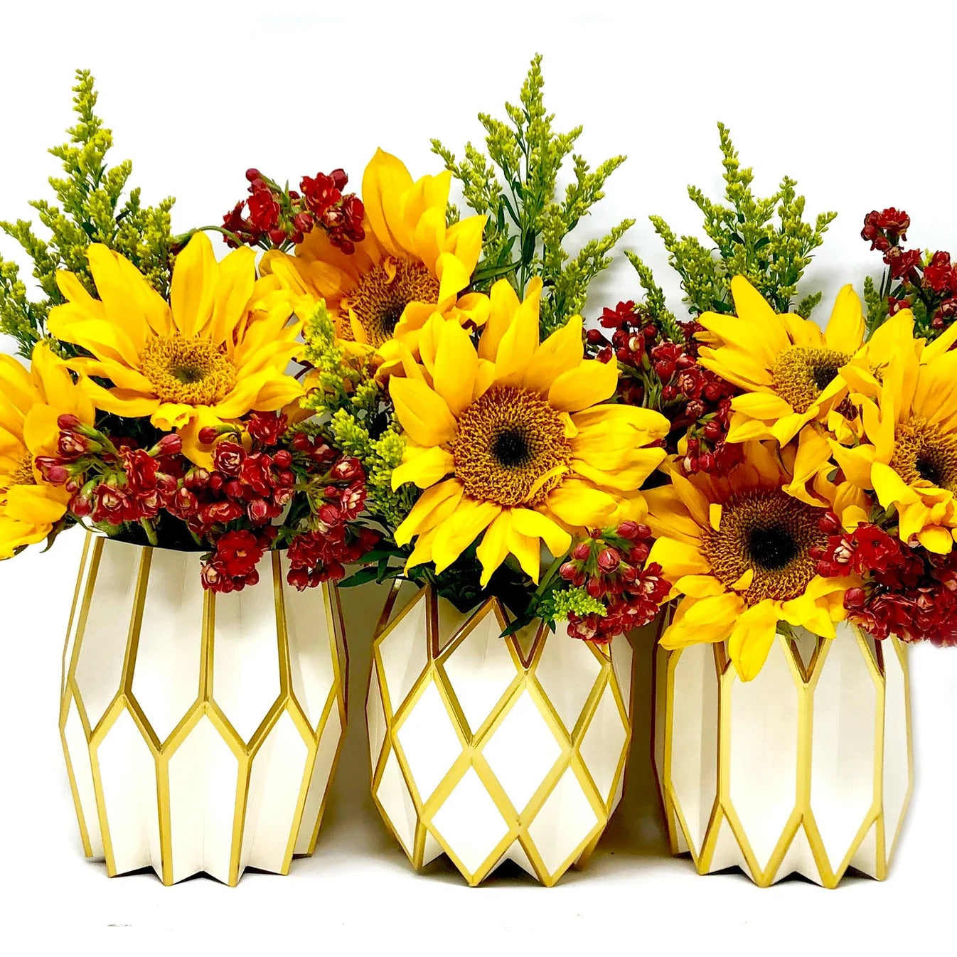 Three Vase Wraps by Lucy Grymes (pearl & gold) from Lucy Grymes hold bright yellow sunflowers, red berries, and green foliage, arranged against a white background.