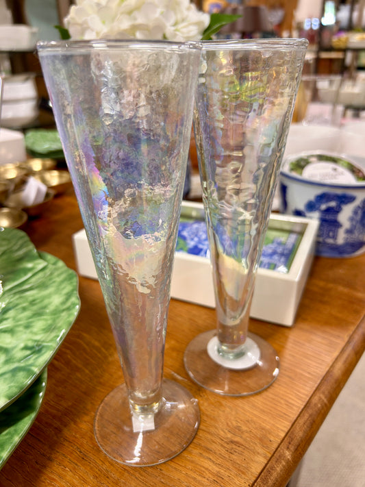 Two Karma "Catalina" champagne flutes with an iridescent, textured glass design sit on a wooden table surrounded by decorative items, including green leaf-shaped plates and a blue-and-white ceramic container in the background.