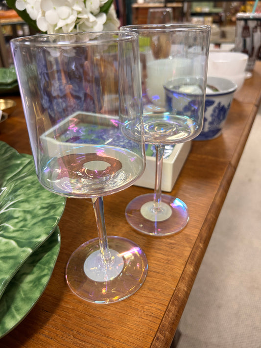 Two Karma "Mid Century" Iridescent Wine Glasses by Karma, featuring delicate iridescent reflections, sit on a wooden table surrounded by green plates, blue-patterned teacups, and other tableware.