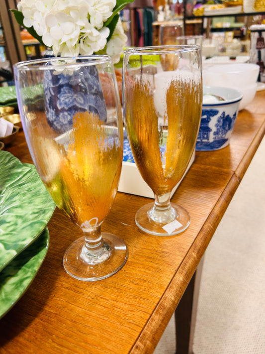 Two ELM Handpainted Gold Water Glasses, with partially worn gold paint and decorated by MS Artist Eileen Morrison, stand on a wooden table among dishes—a green leaf-shaped plate, blue-patterned bowls, and a white floral arrangement in the background.