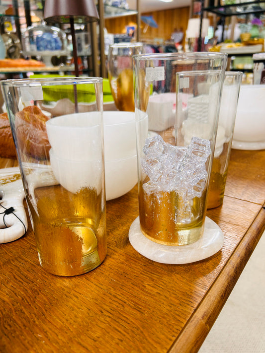Three ELM Handpainted Highball Glasses by ELM, featuring gold accents and designed by Mississippi artist Eileen Morrison, are displayed on a wooden table among white bowls and assorted store items. One glass is filled with clear ice cubes and placed on a round coaster.