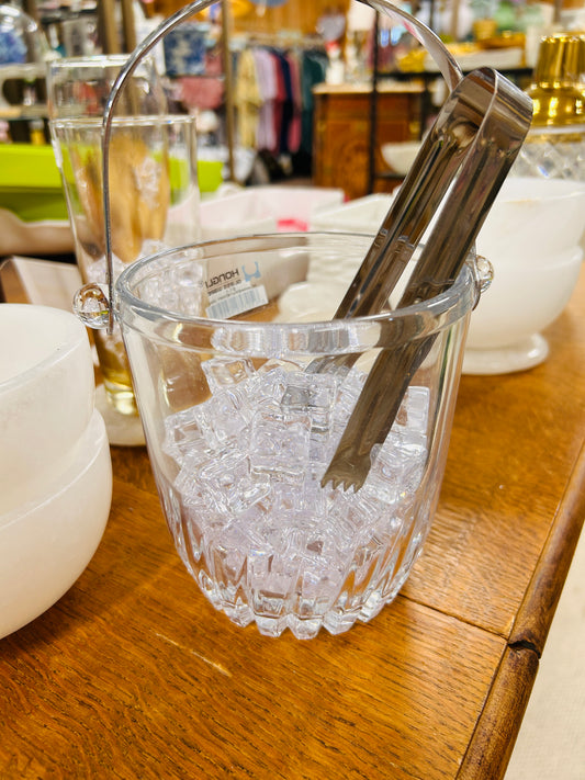 A Hongli Clear Glass Small Ice Bucket filled with acrylic imitation ice cubes and accompanied by metal tongs is displayed on a wooden table among other kitchenware items in the store.