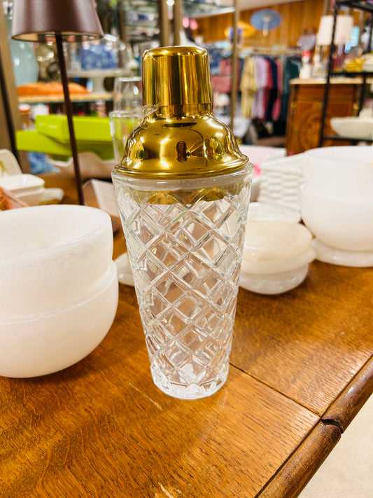 A Cut Glass Cocktail Shaker with a Gold Lid from Holmes Stationers and Gifts sits on a wooden table, surrounded by white bowls, with shelves displaying various items and clothes visible in the background.