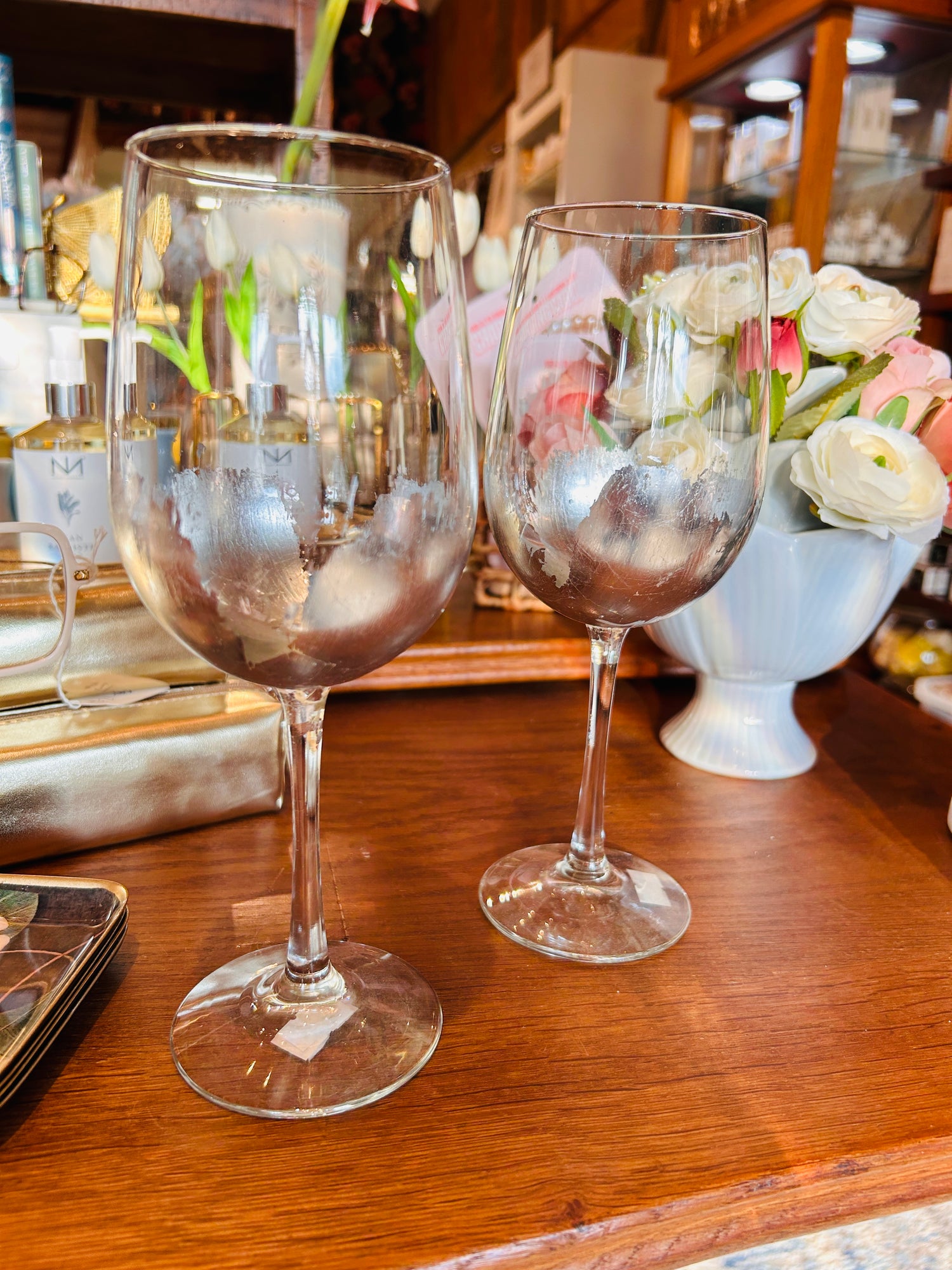 Two partially frosted ELM Handpainted Silver Wine Glasses by ELM are displayed on a wooden surface, with a white vase of flowers and assorted items blurred in the background. Designed by MS artist Eileen Morrison, these glasses add an elegant touch to any setting.