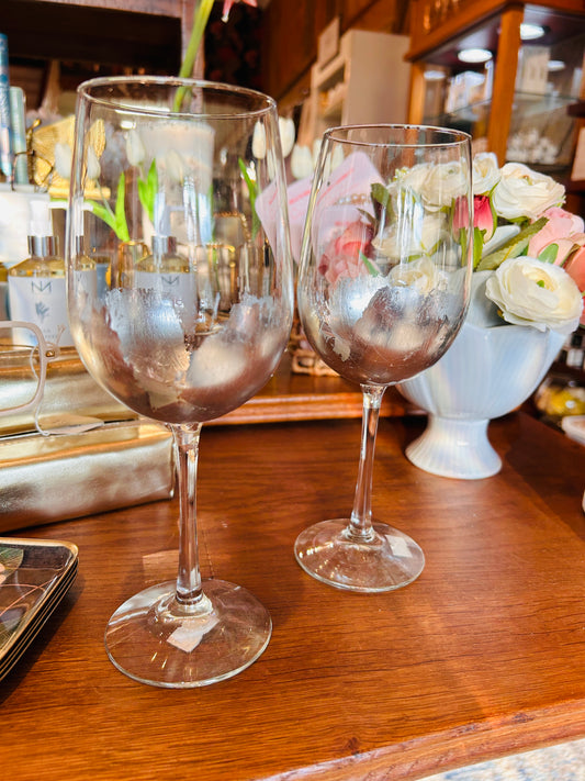 Two partially frosted ELM Handpainted Silver Wine Glasses by ELM are displayed on a wooden surface, with a white vase of flowers and assorted items blurred in the background. Designed by MS artist Eileen Morrison, these glasses add an elegant touch to any setting.