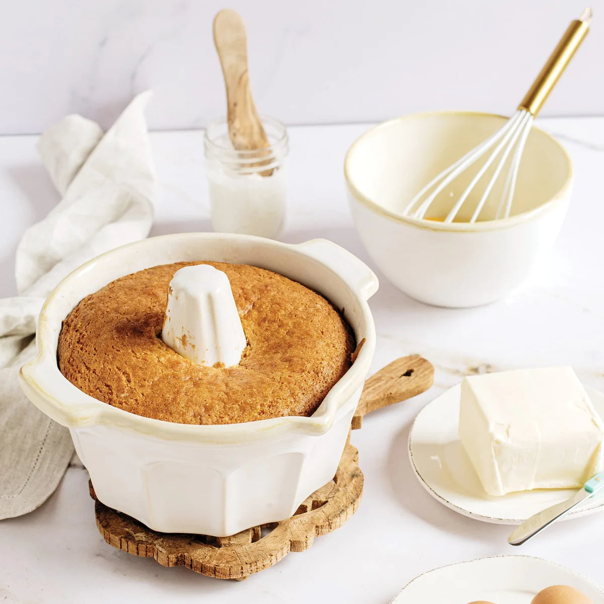 A golden brown cake rests in the Vietri Italian Bakers White Bundt Pan by Vietri, crafted from durable Italian stoneware, on a table alongside a stick of butter, a mixing bowl with a whisk, a jar, and a napkin.