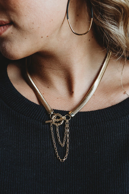 A close-up of a person wearing a gold hoop earring and layered gold necklaces, including the Leslie Curtis Cyrus Necklace by Leslie Curtis—a herringbone gold chain—along with chains featuring toggle closure details, paired with a black ribbed top.