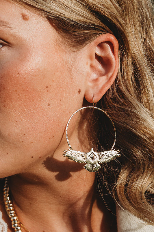 Close-up of a woman's ear wearing the Leslie Curtis Montana Earrings by Leslie Curtis—large, gold hammered hoops featuring an intricate bird design. Her wavy blonde hair frames her sun-kissed skin, enhancing the radiant allure. A glimpse of a gold necklace completes the look, showcasing the elegance of her jewelry care routine.