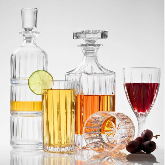 A striking set of 6 includes Godinger "Parallels" Shot Glasses—three crystal shot glasses (two filled with amber and red drinks), a tall glass accented with lime, a wine glass holding red liquid, an empty tumbler resting on its side, and grapes arranged elegantly on a reflective surface.