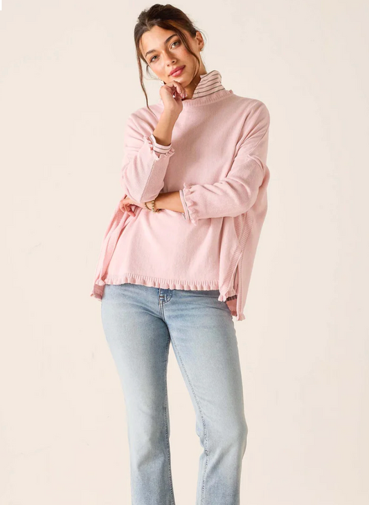 A woman wearing the Mersea Ruffle Catalina Cotton Ruffle Sweater in Rose Quartz and light blue jeans stands against a plain background, with one hand touching her face and the other resting on her hip.