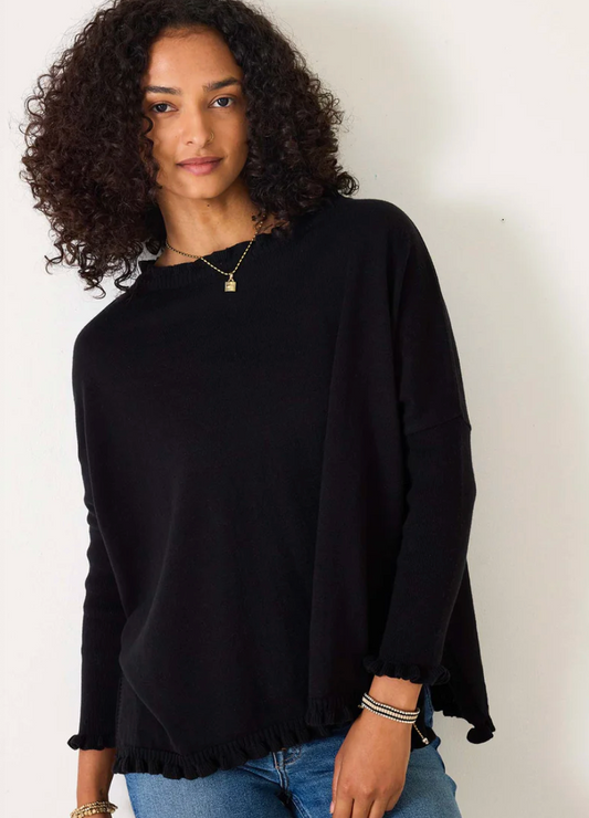 A woman with curly hair is wearing the Mersea Catalina Cotton Ruffle Sweater by Mersea in black, size P, paired with blue jeans. She stands against a white wall and looks at the camera.