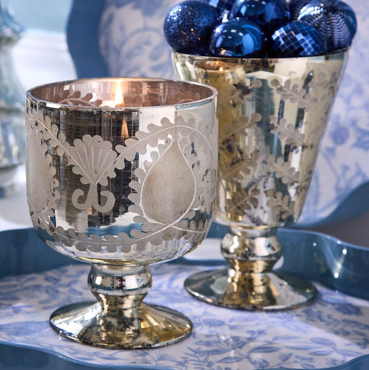Two’s Company Etched Mercury Glass Candleholders, with their vintage-inspired silver detailing, rest on a blue and white tray—one holds a lit candle while the other contains shiny blue ornaments.
