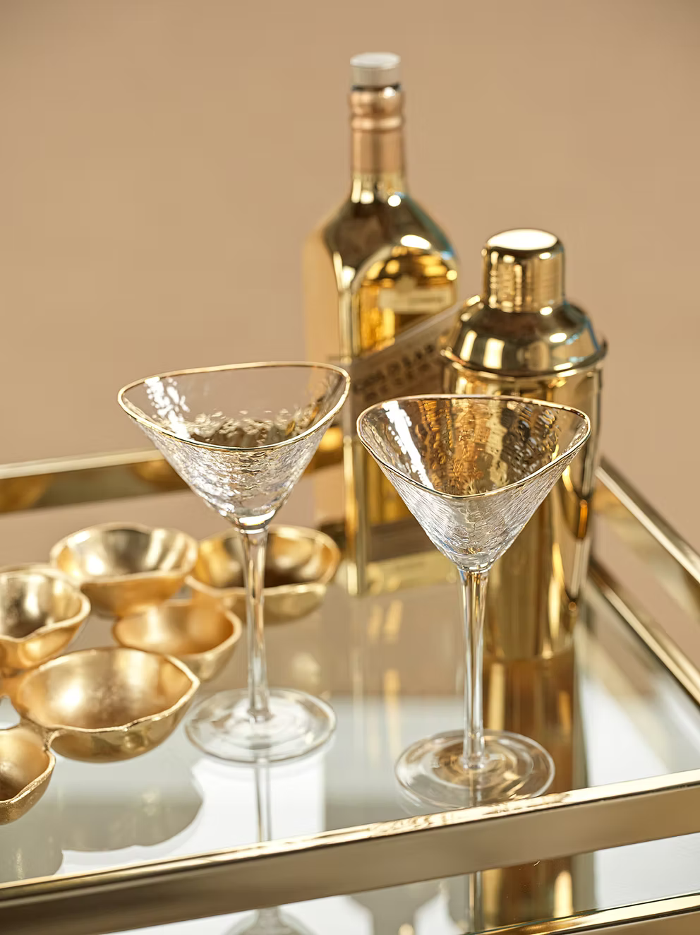 Two Zodax "Kampari" martini glasses rest on a mirrored tray, accompanied by a gold cocktail shaker, a gold bottle, and small gold bowls, creating a luxurious and elegant bar setup.