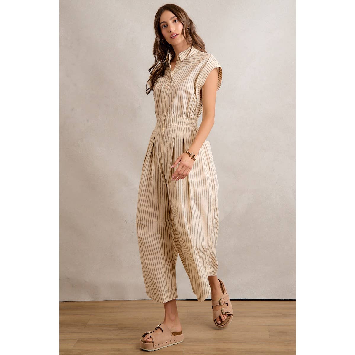 A woman models the So Me - SLEEVELESS WIDE LEG STRIPE JUMPSUIT, featuring beige and white stripes, a collared neckline, and wide legs. She pairs it with tan platform sandals and stands on a wooden floor against a neutral background.