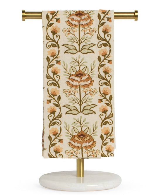 A brown and orange floral-patterned fabric is elegantly draped over a gold and marble stand against a white background, highlighting The Royal Standard’s Adeline Hand Towel as both a luxurious, soft essential and a stylish decorative accent.
