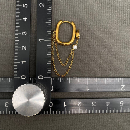 A pair of Mio Queena - Chain Fringe Zircon Inlaid 18K Gold Plated SS Earrings - FGS rests on a dark textured surface, with two delicate chains and a small gemstone accent. Crossed black rulers marked in centimeters measure the earrings. Crafted by Mio Queena, these earrings feature durable 18K gold plating and are designed for a non-tarnish finish, ensuring lasting shine.
