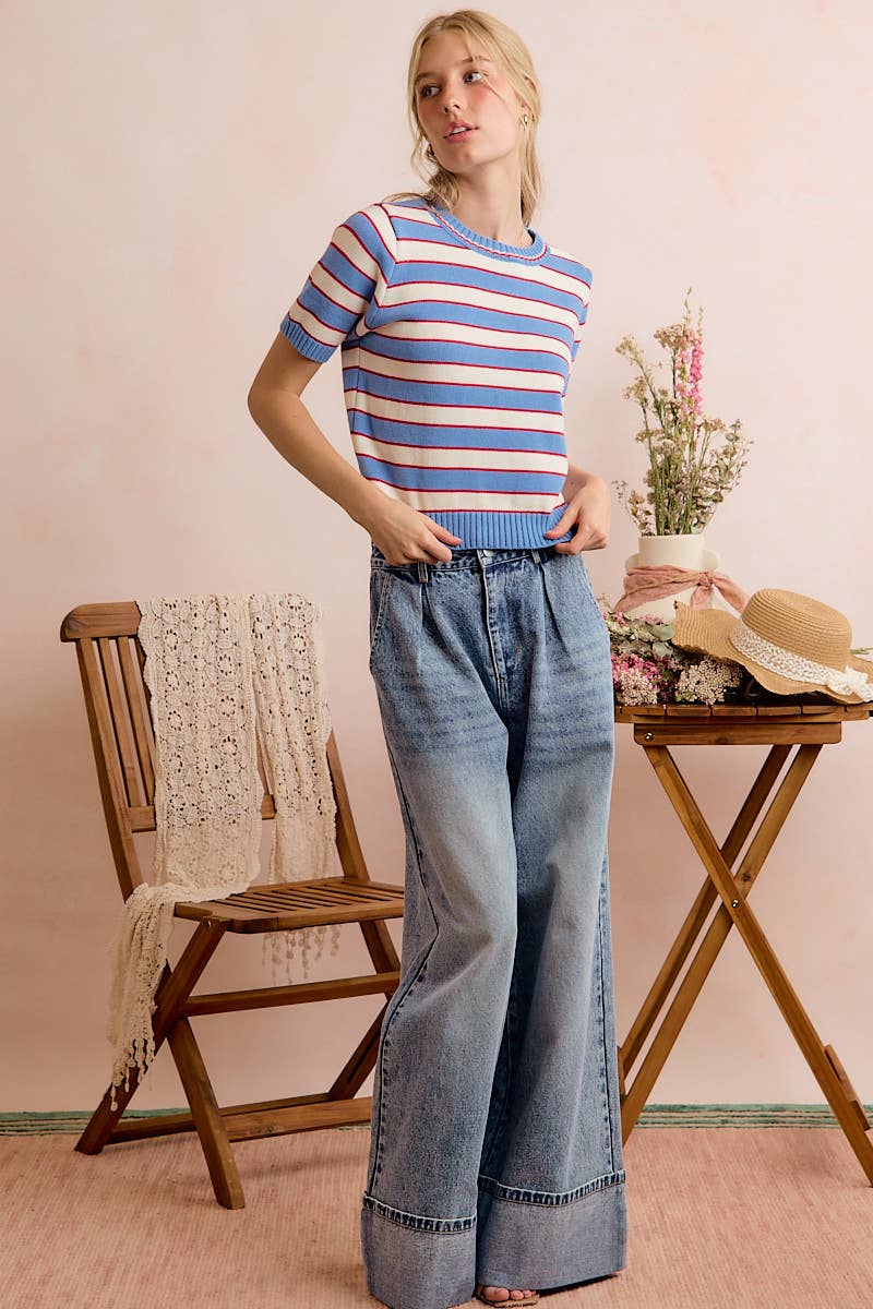 A woman stands indoors wearing the So Me STRIPE KNIT CREW NECK SHORT SLEEVES SWEATER TOP paired with wide-leg jeans. She is next to a wooden chair and table adorned with flowers, a lace cloth, and a straw hat, set against a light pink background.