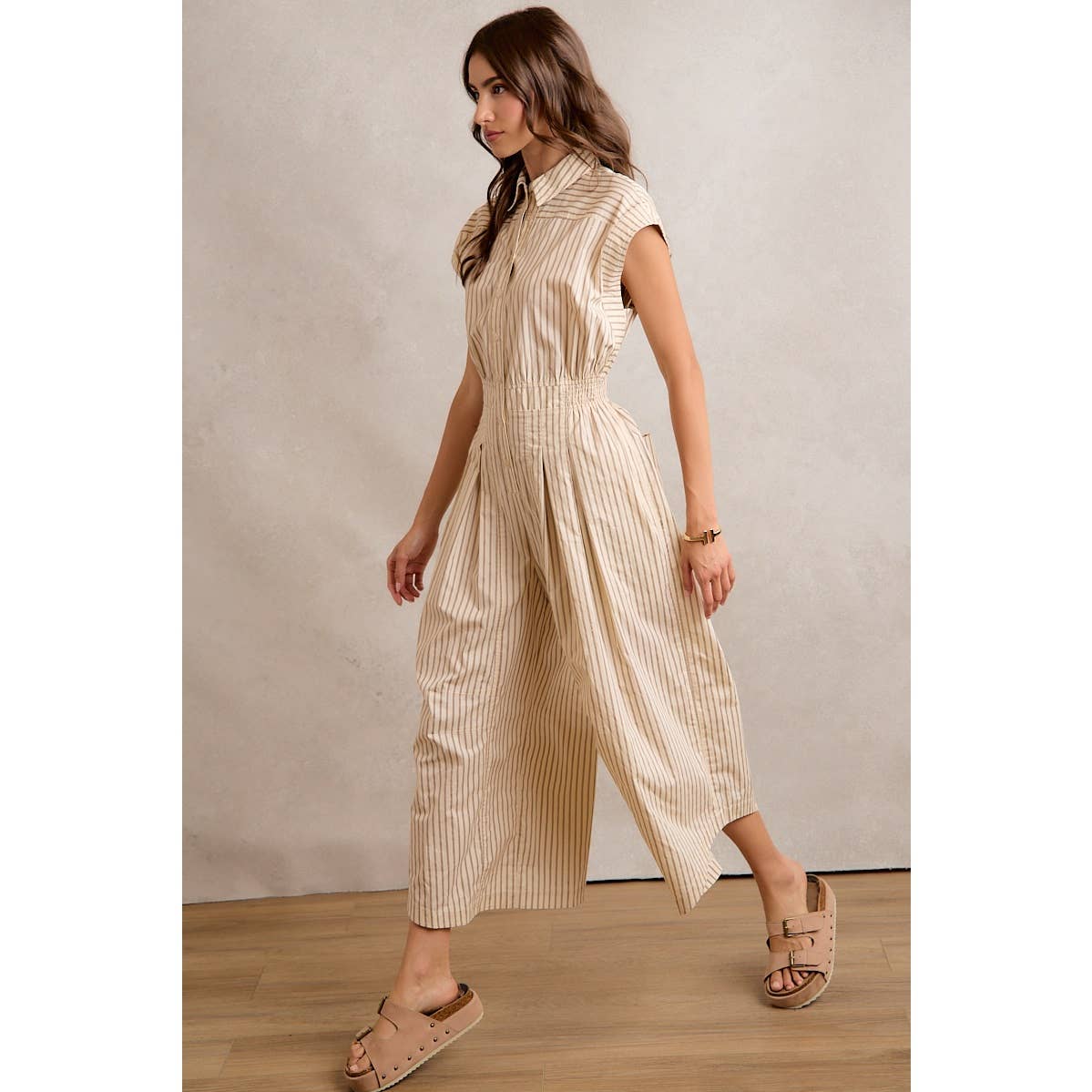 A woman in the So Me SLEEVELESS WIDE LEG STRIPE JUMPSUIT walks indoors on a wooden floor, wearing tan sandals and a bracelet, her long brown hair styled in loose waves.