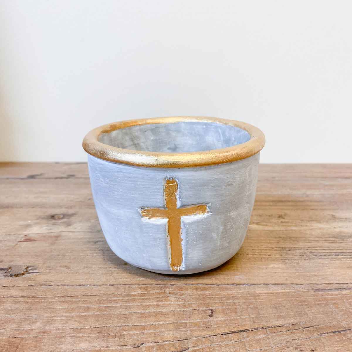 The Royal Standard Stockholm Cross Candle Holder by The Royal Standard, a small round gray pot featuring a gold rim and a gold cross on the front, sits on a wooden surface against a plain background.
