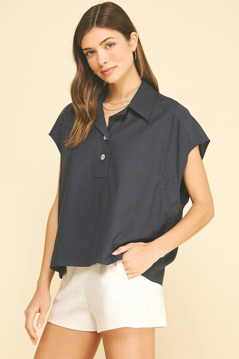 A woman wearing the PINCH - SOLID CAP SLEEVE BLOUSE TOP by PINCH, a loose-fitting navy blouse with a classic collar and white buttons, paired with white shorts, stands against a light beige background.