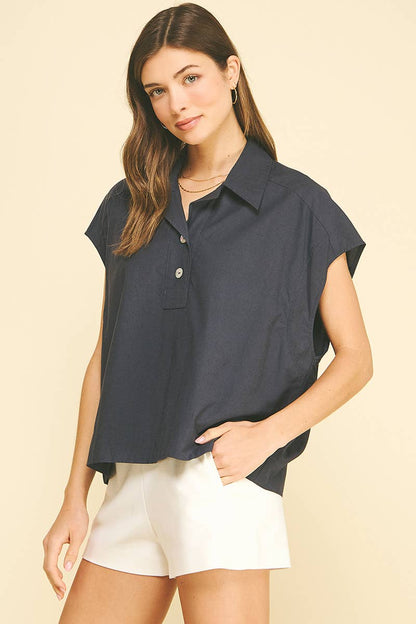 A woman wearing the PINCH - SOLID CAP SLEEVE BLOUSE TOP by PINCH, a loose-fitting navy blouse with a classic collar and white buttons, paired with white shorts, stands against a light beige background.
