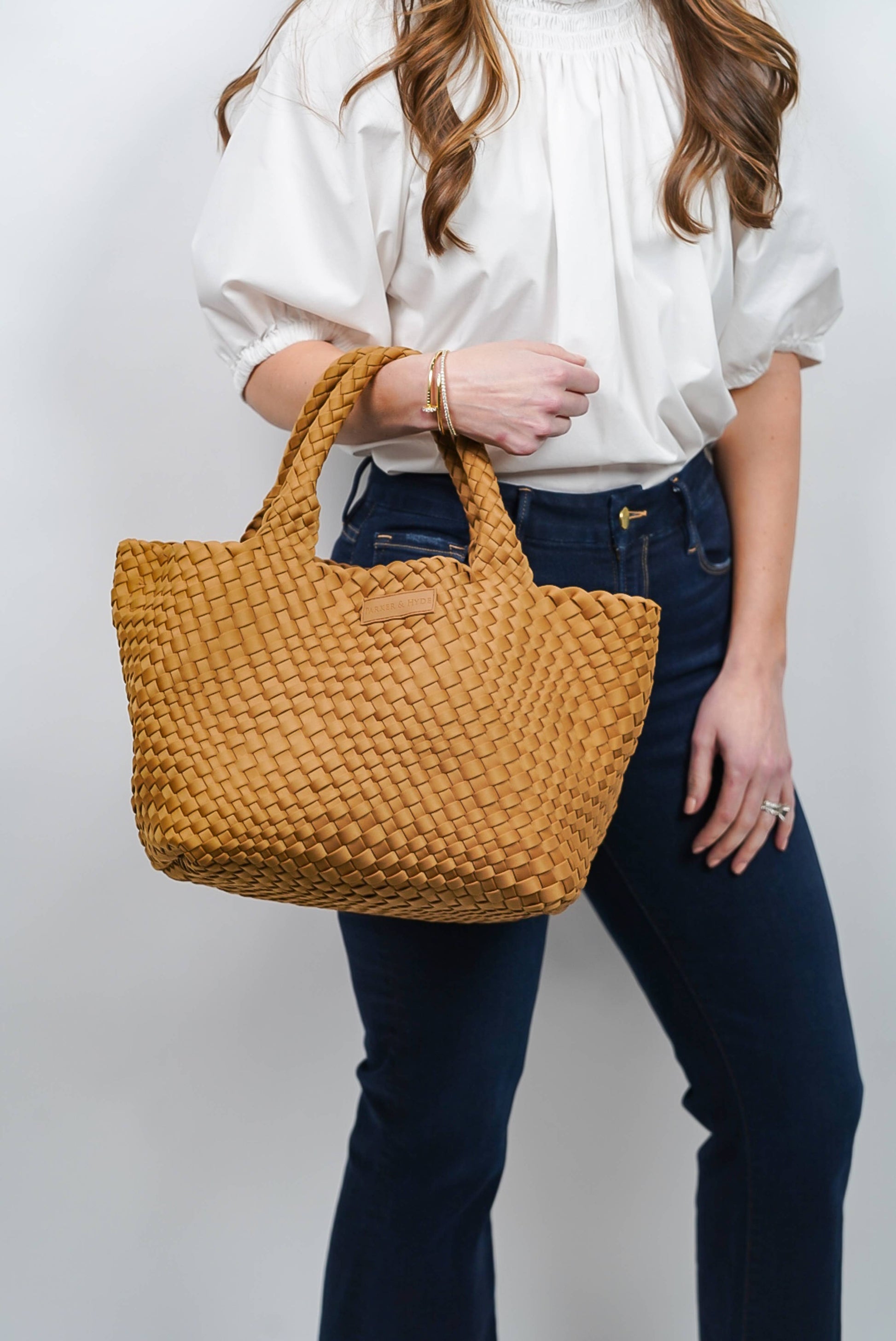 A woman, seen from the torso down and accessorized with bracelets and a ring, wears a white blouse and dark jeans while holding the Parker & Hyde Classic Woven Tote in Camel.